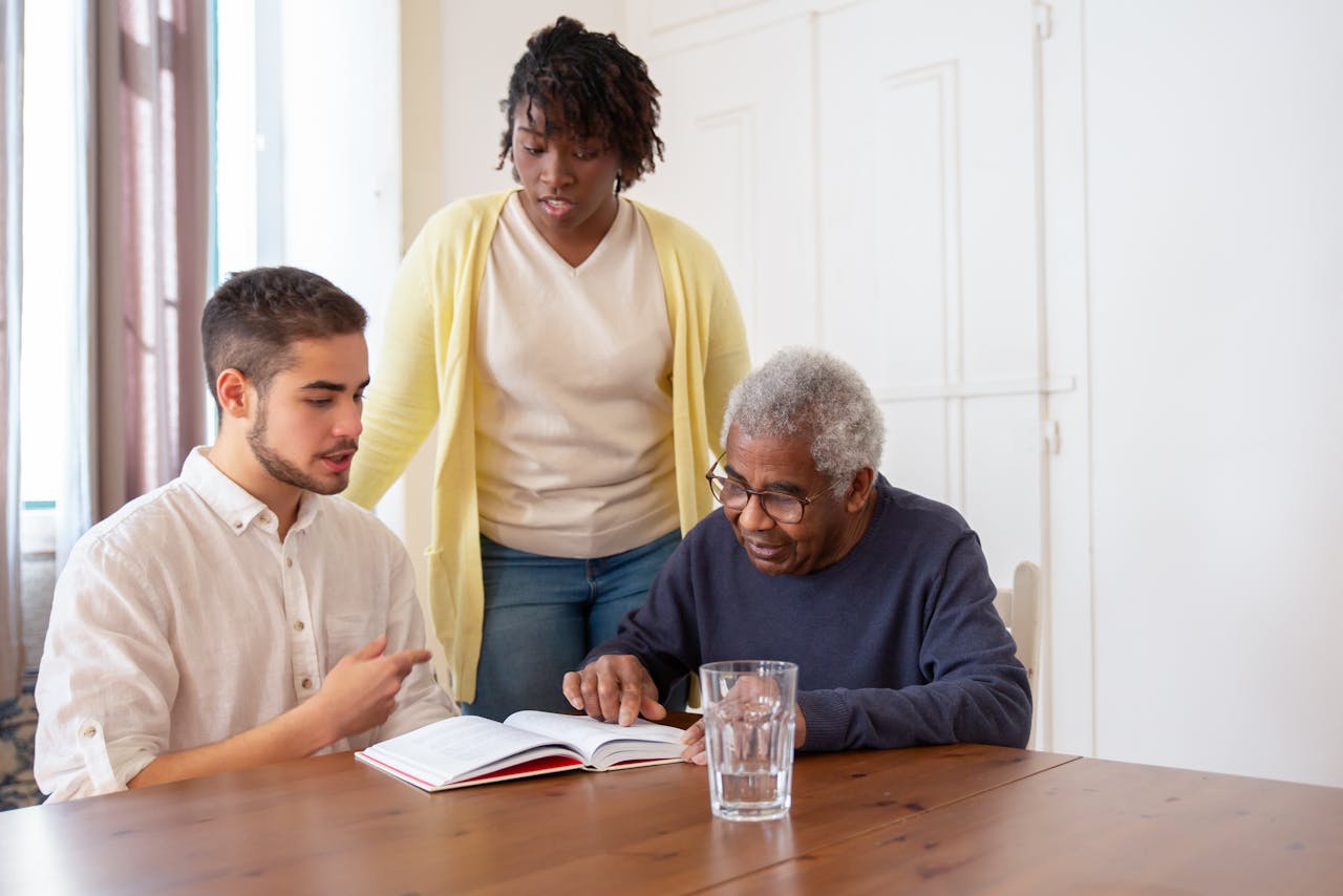Young adults assisting an elderly man with reading in a nursing home.